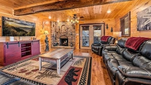 L’intérieur d’une cabane en rondins, avec une cheminée en pierre, un canapé en cuir et un meuble de rangement en bois pour les appareils multimédias.