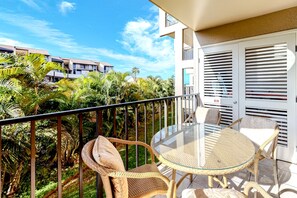 A balcony with a glass table and wicker chairs, overlooking a lush garden and neighboring buildings. - *NEW* Fall Deal Kamaole Beach III Retreat With Pool, Hot Tub and Tennis (Kihei)