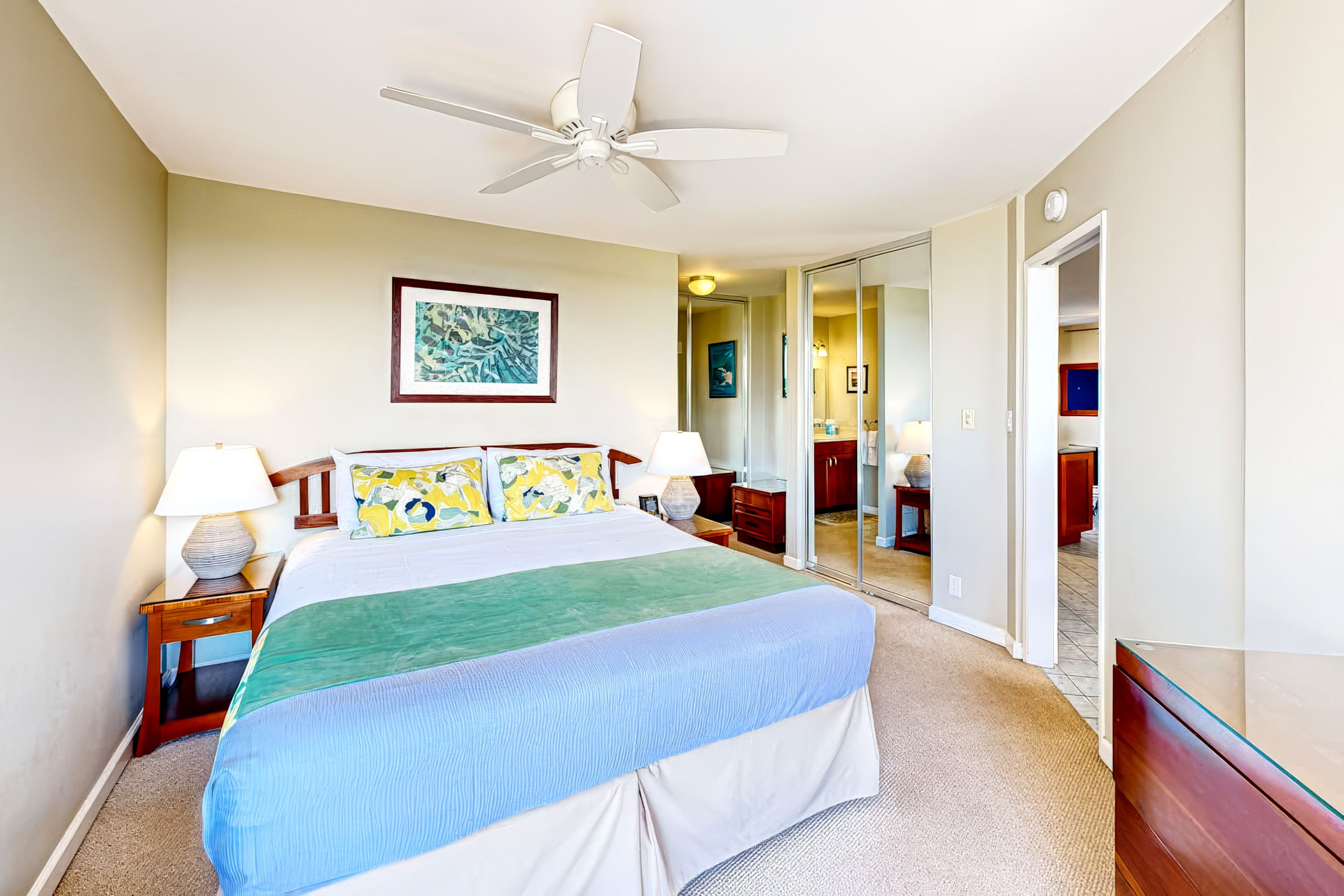 A hotel room with a bed, a ceiling fan, a large mirror, and artwork on the wall.