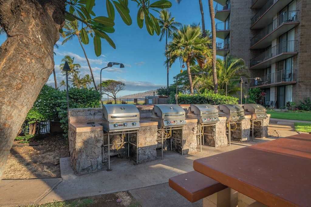 An outdoor barbecue area with stone grills, a picnic table, and a view of a building with balconies and palm trees.