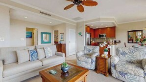 Sala de estar con sofá, sillones, una mesa de centro, ventilador de techo y una zona de cocina con armarios y electrodomésticos.