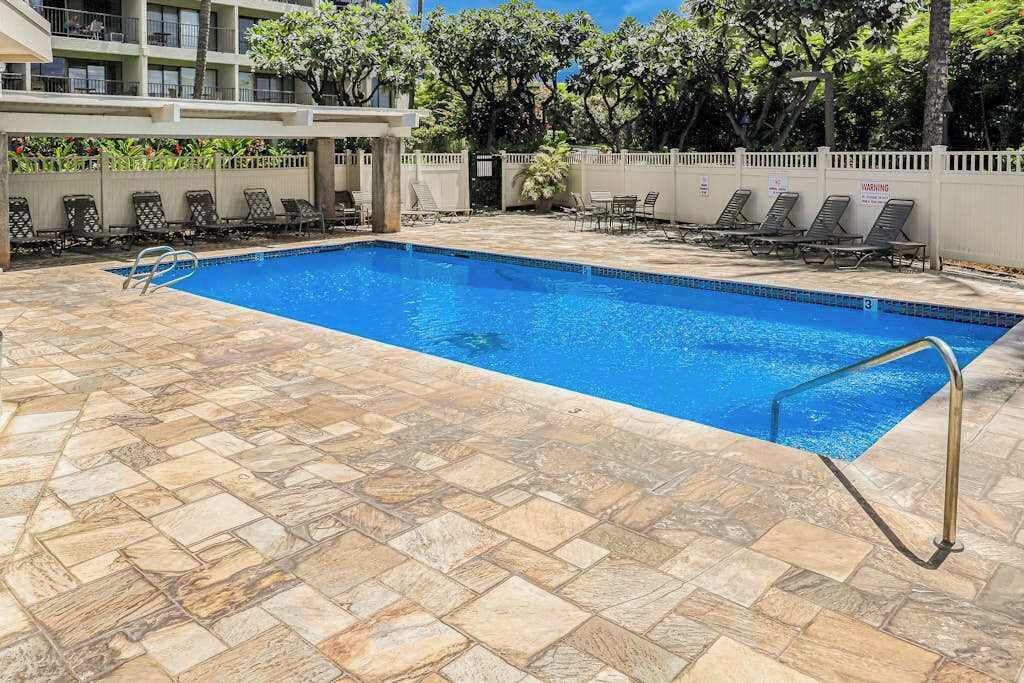 A rectangular swimming pool with a tiled deck, surrounded by lounge chairs and a white fence.