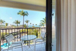 Ein Balkon mit Glastisch und zwei Stühlen, mit Blick auf einen Strand mit Palmen und einem klaren Himmel.