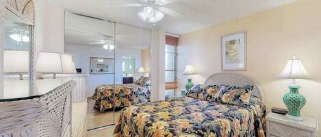 Un dormitorio con cama, ventilador de techo, mesita de noche, lámpara, espejo y ventana.