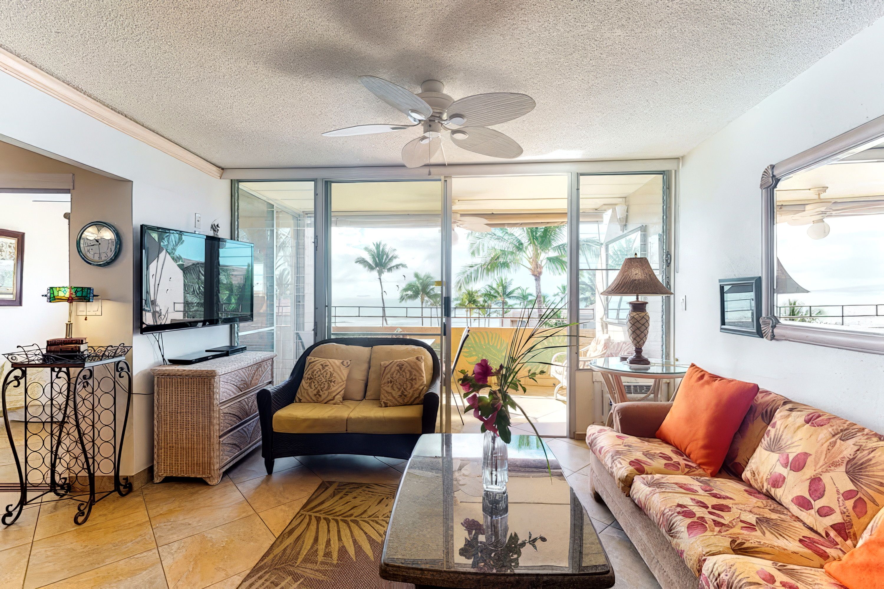A living room with a ceiling fan, a sofa, a coffee table, a TV, and a view of palm trees through large windows.