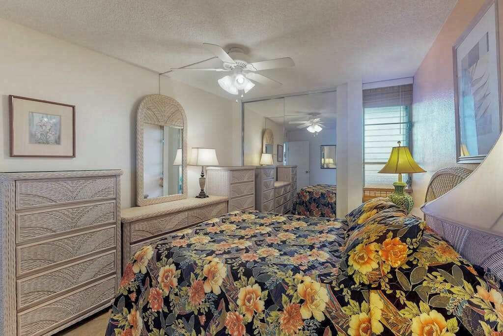 Une chambre à coucher comprenant un lit, une commode, un miroir, un ventilateur de plafond et une fenêtre avec des stores.