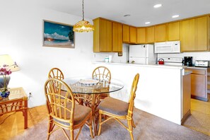 A compact kitchen with wooden cabinets, a dining area with a glass table and wicker chairs, and a painting on the wall. - *New* Fall $pecial Garden-View Kihei Condo for Four Near Kamaole III (Kihei)