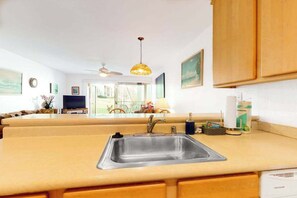 A kitchen with a stainless steel sink, wooden cabinets, a ceiling fan, and a view of the living room. - *New* Fall $pecial Garden-View Kihei Condo for Four Near Kamaole III (Kihei)