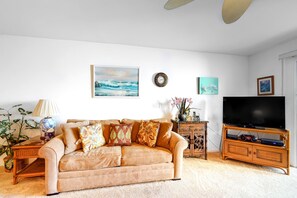 A living room with a beige sofa, a flat-screen TV, a wooden cabinet, and a wall clock. - *New* Fall $pecial Garden-View Kihei Condo for Four Near Kamaole III (Kihei)