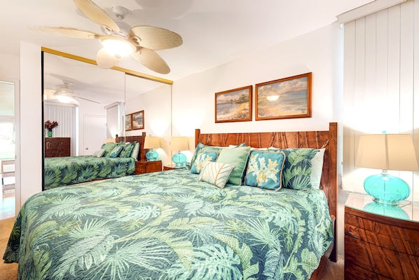 A bedroom with a large bed, a ceiling fan, two wall-mounted paintings, and a nightstand with a lamp. - *New* Fall $pecial Garden-View Kihei Condo for Four Near Kamaole III (Kihei)