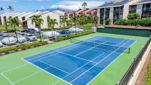 Un court de tennis avec un filet, entouré d’une clôture, et situé à côté d’un parking abrité.