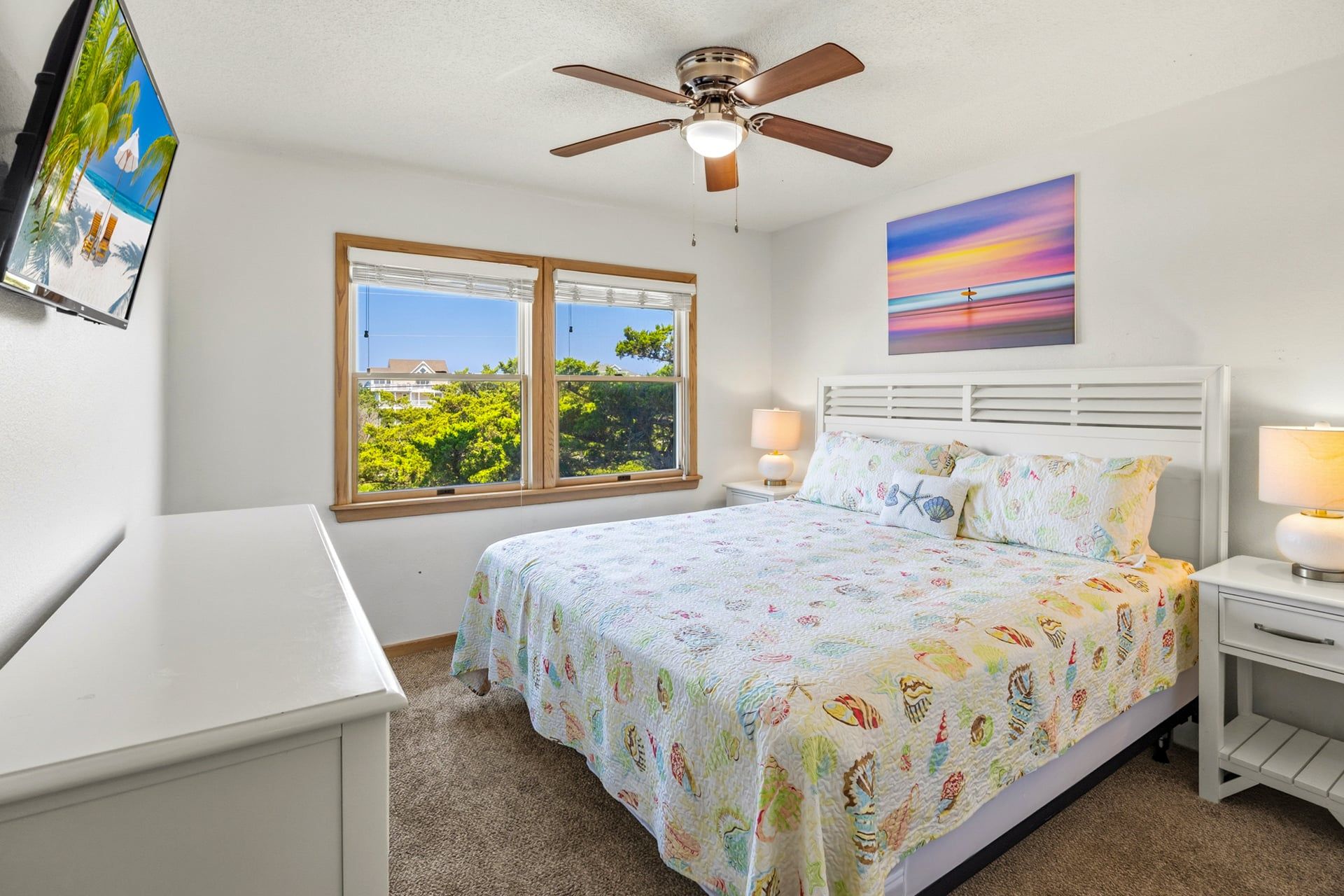A bedroom with a bed, two bedside tables, a ceiling fan, a window with a view, and a painting on the wall.