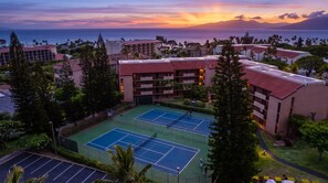 Un court de tennis entouré de bâtiments d’hôtel, avec vue sur l’océan au coucher du soleil.