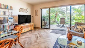 Sala de estar con televisor, estanterías, comedor y puerta corrediza de vidrio a un balcón con mobiliario exterior.