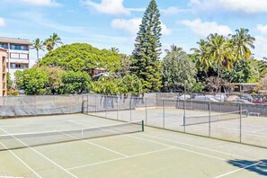 An outdoor tennis court surrounded by a fence, with a backdrop of trees and buildings. - New Fall Deal Top-Floor Ocean Views Across from Charley Young Beach (Kihei)