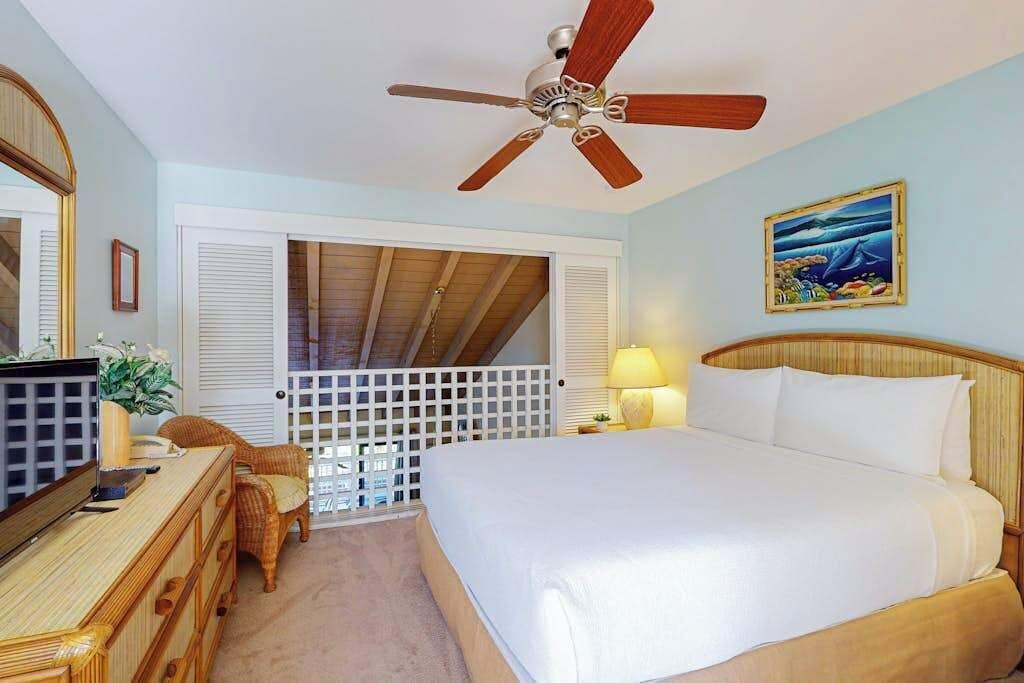Un dormitorio con cama, ventilador de techo, televisor, cómoda, silla, lámpara y un cuadro en la pared.