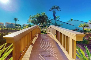Eine Holzbrücke führt zu einem Gebäude mit modernem Design, umgeben von Grünpflanzen und Palmen.