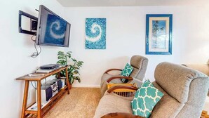 Ein Wohnzimmer mit einer Couch, einem Fernseher, einem Holztisch mit Pflanze und Kunstwerken an der Wand.