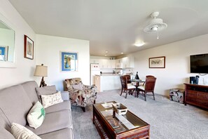 Een woonkamer met een bank, fauteuil, salontafel en een eethoek met stoelen en een tafel. Achtergrond is een keuken met kasten en een magnetron.