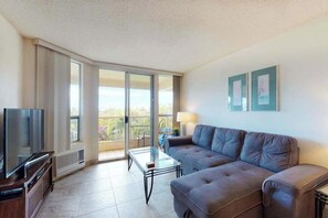 A modern living room with a flat-screen TV, a sofa, a glass coffee table, and a balcony with a view. - NEW Fall Special Fourth-Floor Kihei Condo with Ocean Glimpse and Beach Access (Kihei)