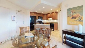 A modern kitchen with wooden cabinets, a central island, and a dining area with a glass table and wicker chairs. - NEW Fall Special Fourth-Floor Kihei Condo with Ocean Glimpse and Beach Access (Kihei)