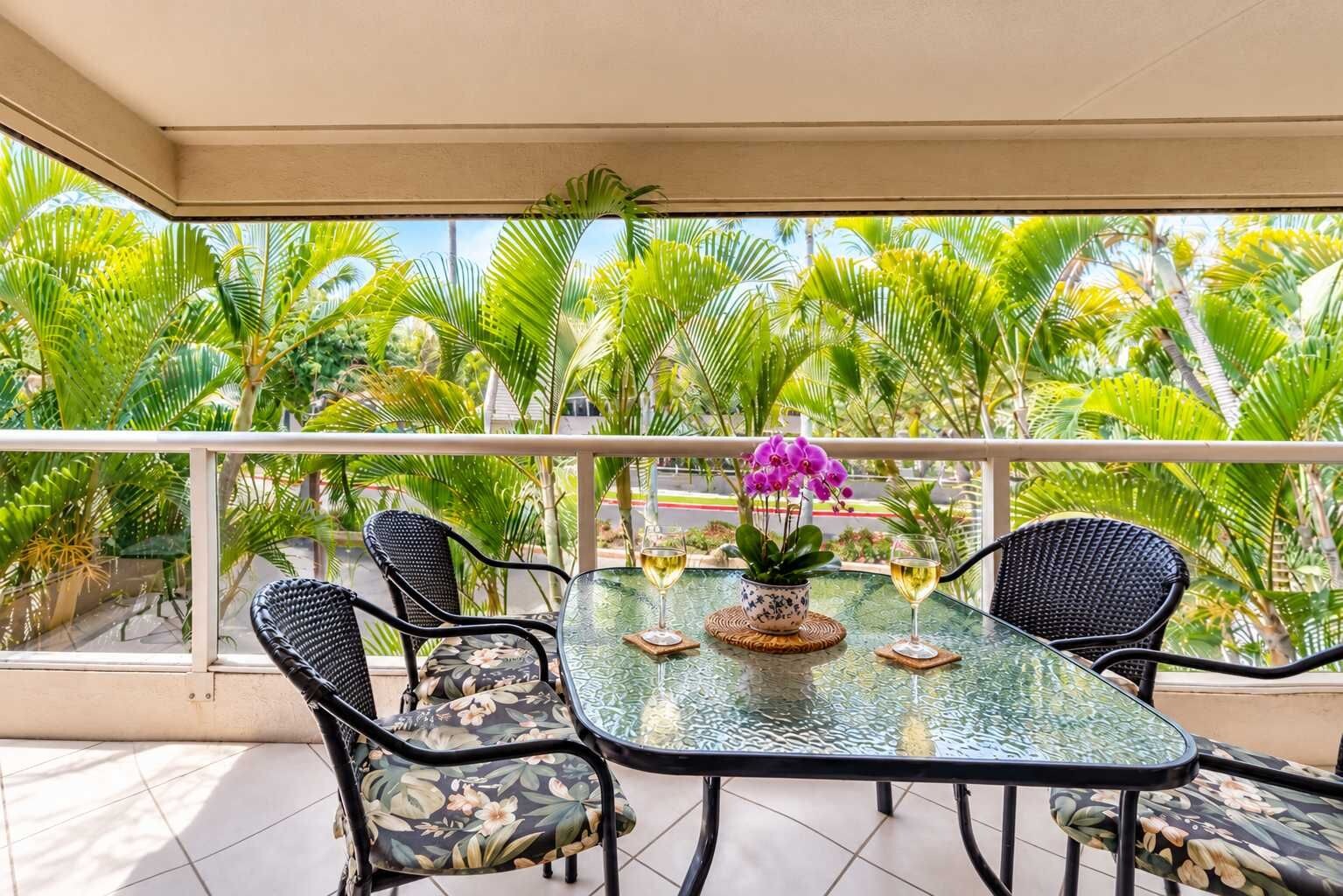 A glass-top table with chairs, a potted orchid, and two wine glasses on a balcony with a view of palm trees.