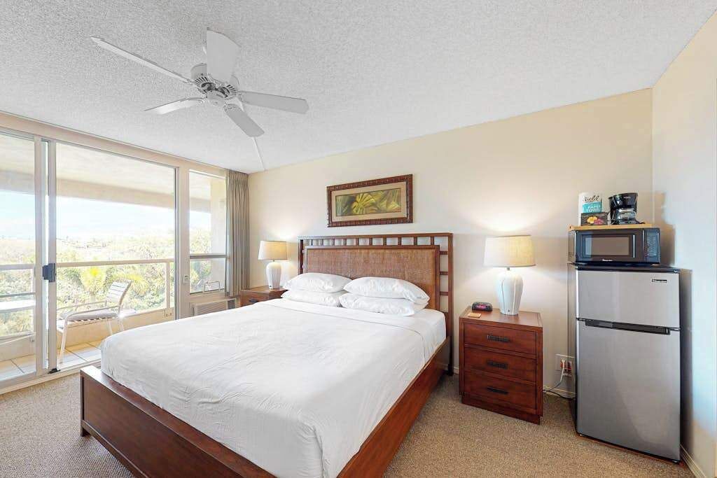 Una camera d'albergo con un letto, una ventola a soffitto, un frigorifero, un comodino, una lampada e una finestra vista.