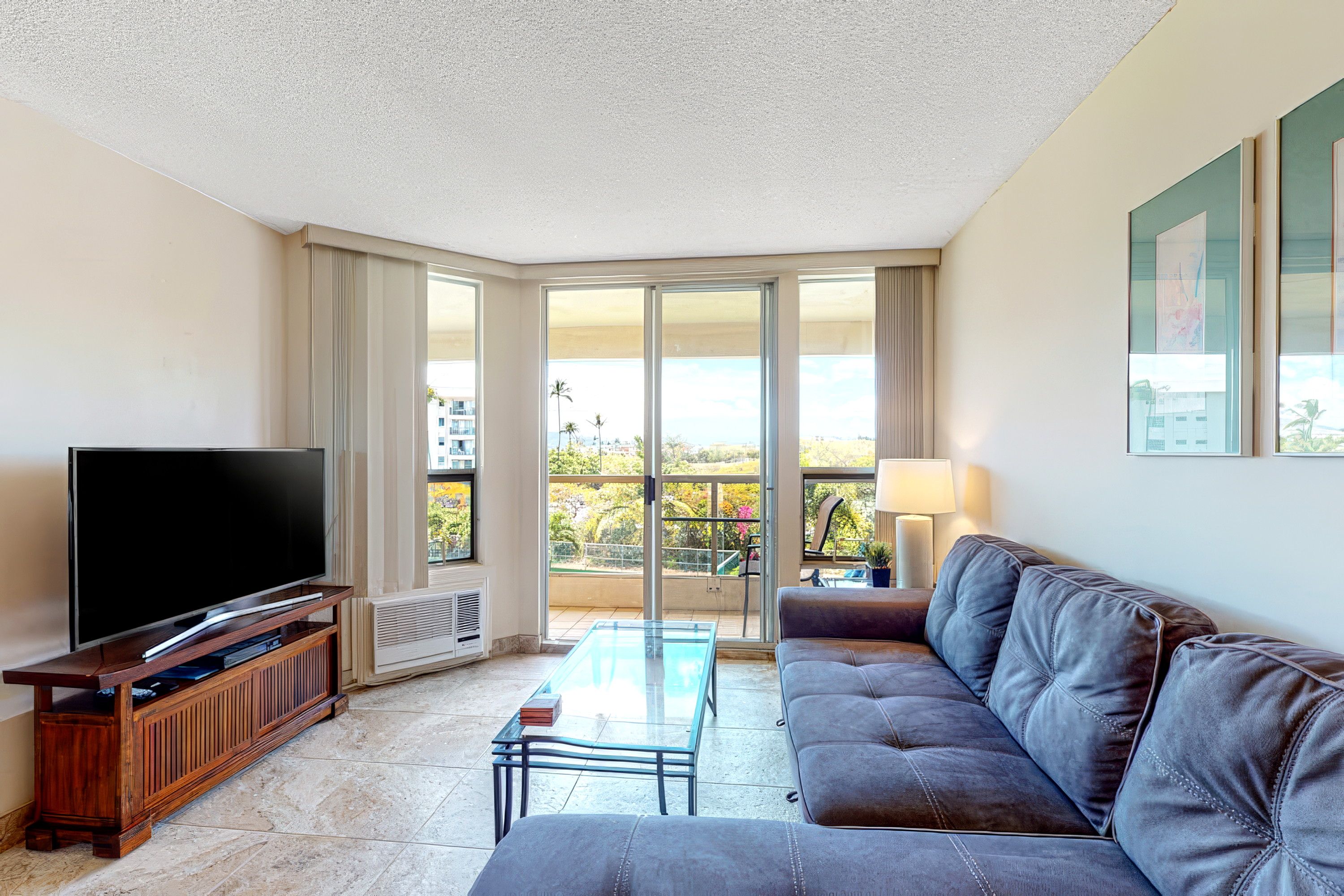 A living room with a flat-screen TV, a sofa, a coffee table, and a balcony with a view of buildings and trees.