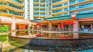 Ein mehrstöckiges Hotel mit einem zentralen Innenhof, der einen spiegelnden Wasserspielplatz, Sitzgelegenheiten im Freien und einen überdachten Bereich mit Sonnenschirmen bietet.