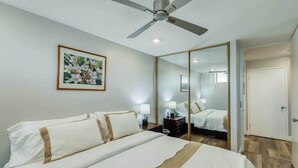 A hotel room with a bed, a ceiling fan, a bedside table with a lamp, a large mirror, and a door. - *New* Fall Deal Panoramic Kahana Oceanfront for Two (Lahaina)
