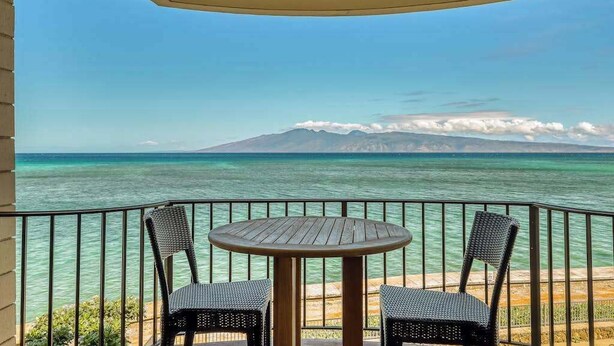 A balcony with two wicker chairs and a round table, overlooking a clear blue sea and a mountain in the distance. - *New* Fall Deal Panoramic Kahana Oceanfront for Two (Lahaina)