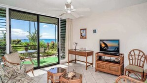 Una habitación con un amplio ventanal corredera que da a un balcón, un ventilador de techo, televisión, muebles de mimbre y vista al océano.