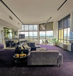 A modern living room with a large sofa, a coffee table, and a dining area with bar stools. - Gold Coast Penthouse Retreat - Where every window is a view! (Surfers Paradise)