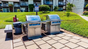 An outdoor barbecue area with a grill and a trash can, located in a landscaped garden with a building in the background. - *New* Fall $ale Kamaole III Beach Days with a Kihei Lanai (Kihei)
