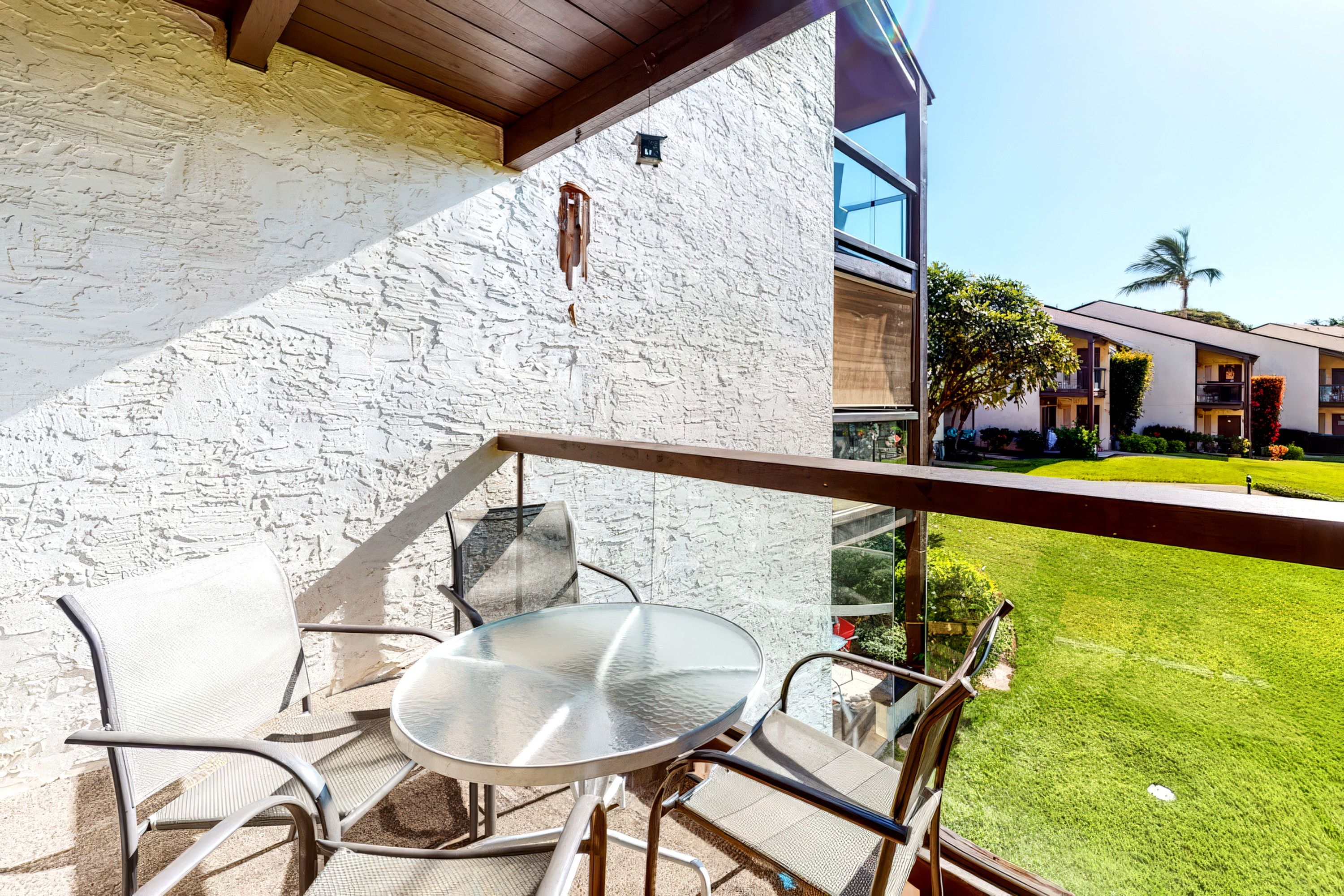 A balcony with a glass table and chairs, overlooking a well-maintained lawn and a building with a white exterior.