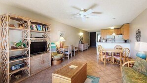 Una sala de estar con televisión, muebles de mimbre, un comedor y una cocina al fondo.