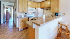 Una cocina con armarios de madera, microondas, refrigerador, encimera y un taburete.