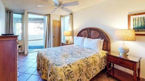 Una habitación de hotel con cama, ventilador de techo, mesita de noche con lámpara y ventana con cortinas.