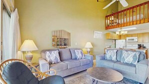 Un salon avec un canapé, un fauteuil et une table basse, une cuisine à l’arrière-plan et un balcon avec une balustrade.