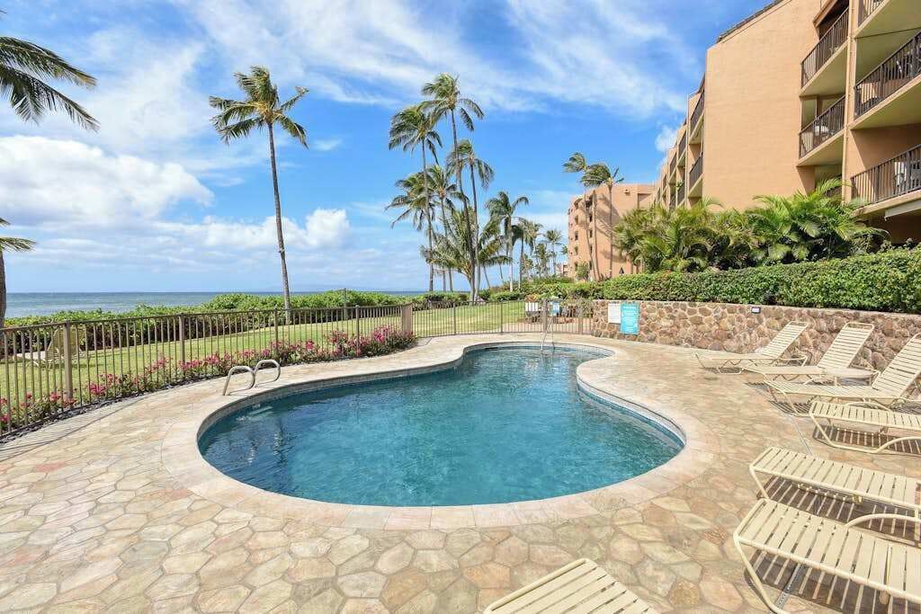 A swimming pool with a curved edge, surrounded by lounge chairs and palm trees, with a building in the background.