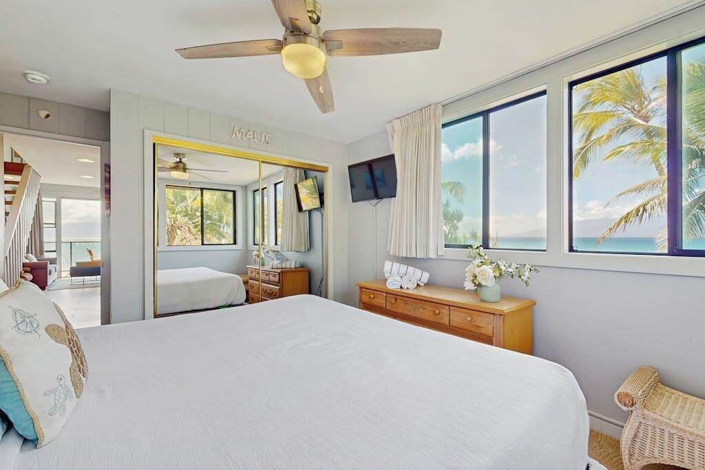 Une chambre avec un grand lit, un ventilateur de plafond, une télévision, une commode et une fenêtre donnant sur des palmiers.