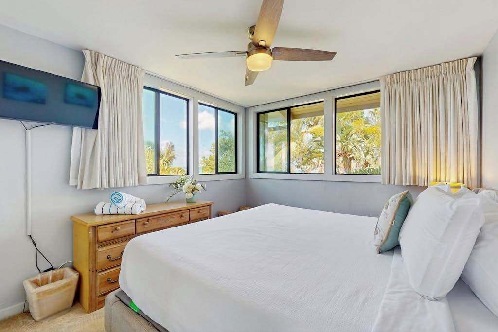 Quarto com cama, ventilador de teto, TV na parede e janelas com cortinas.