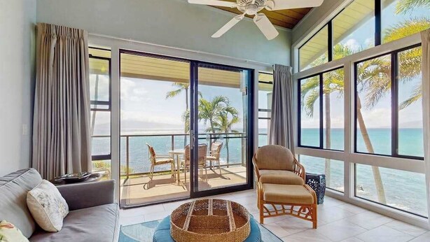 Ein Zimmer mit einer Couch, einem Sessel, einem Tisch und Blick auf das Meer durch große Fenster.