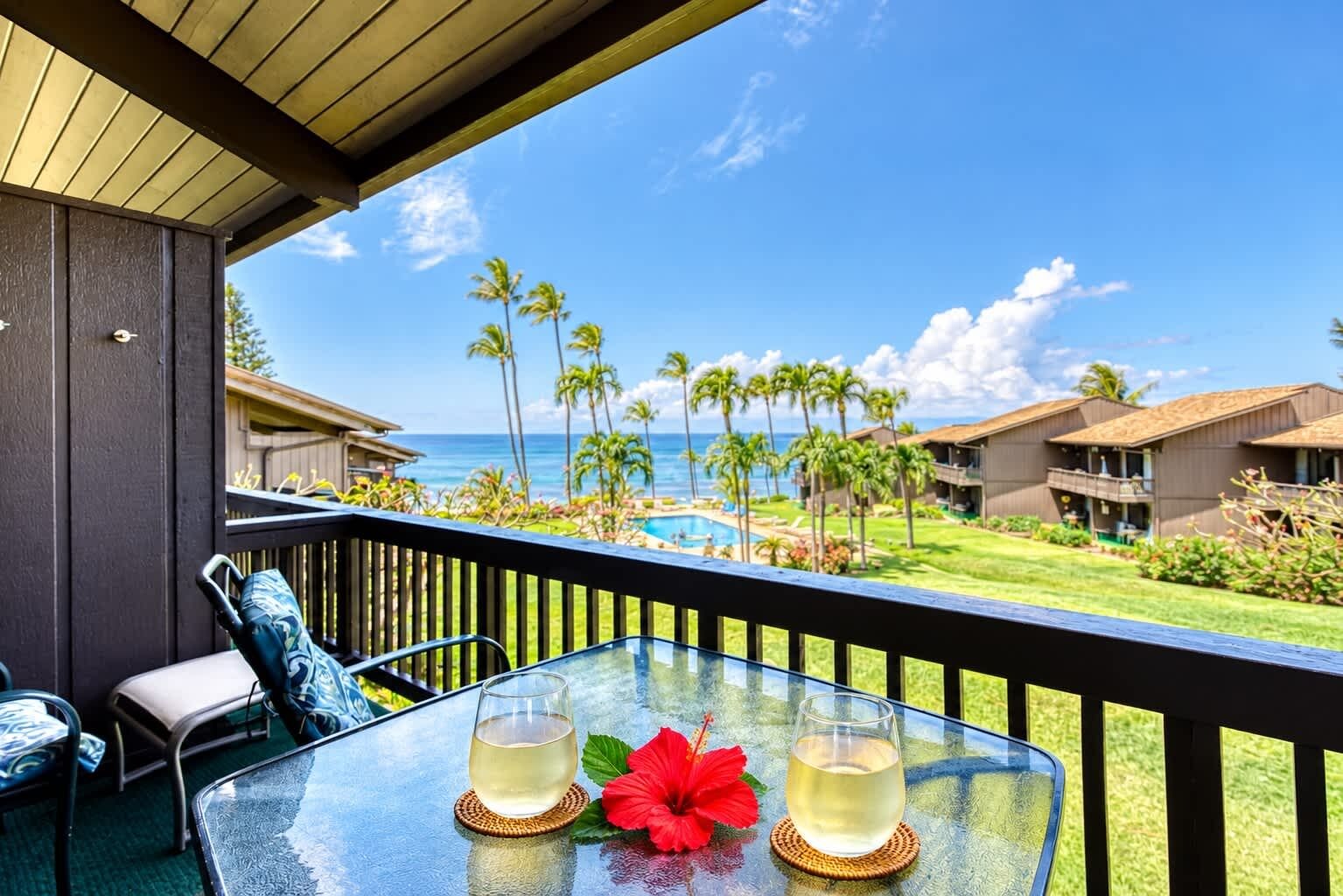 A balcony with a glass table, two glasses of a clear beverage, and a red flower.