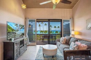 A living room with a TV, sofa, ottoman, and a balcony with a view of the ocean and palm trees. - Stunning Ocean View 2 Bedroom Condo in Lahaina (Lahaina)