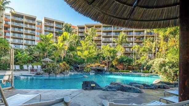 Uma área de piscina de hotel com espreguiçadeiras, um elemento aquático e uma estrutura coberta de palha.
