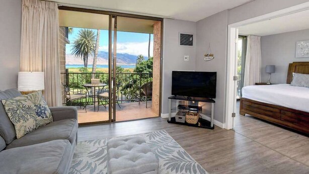A hotel room with a balcony, a sofa, a TV, a bed, and a view of the outdoors. - New Oceanfront Kihei Condo with Pool and Hot Tub (Kihei)