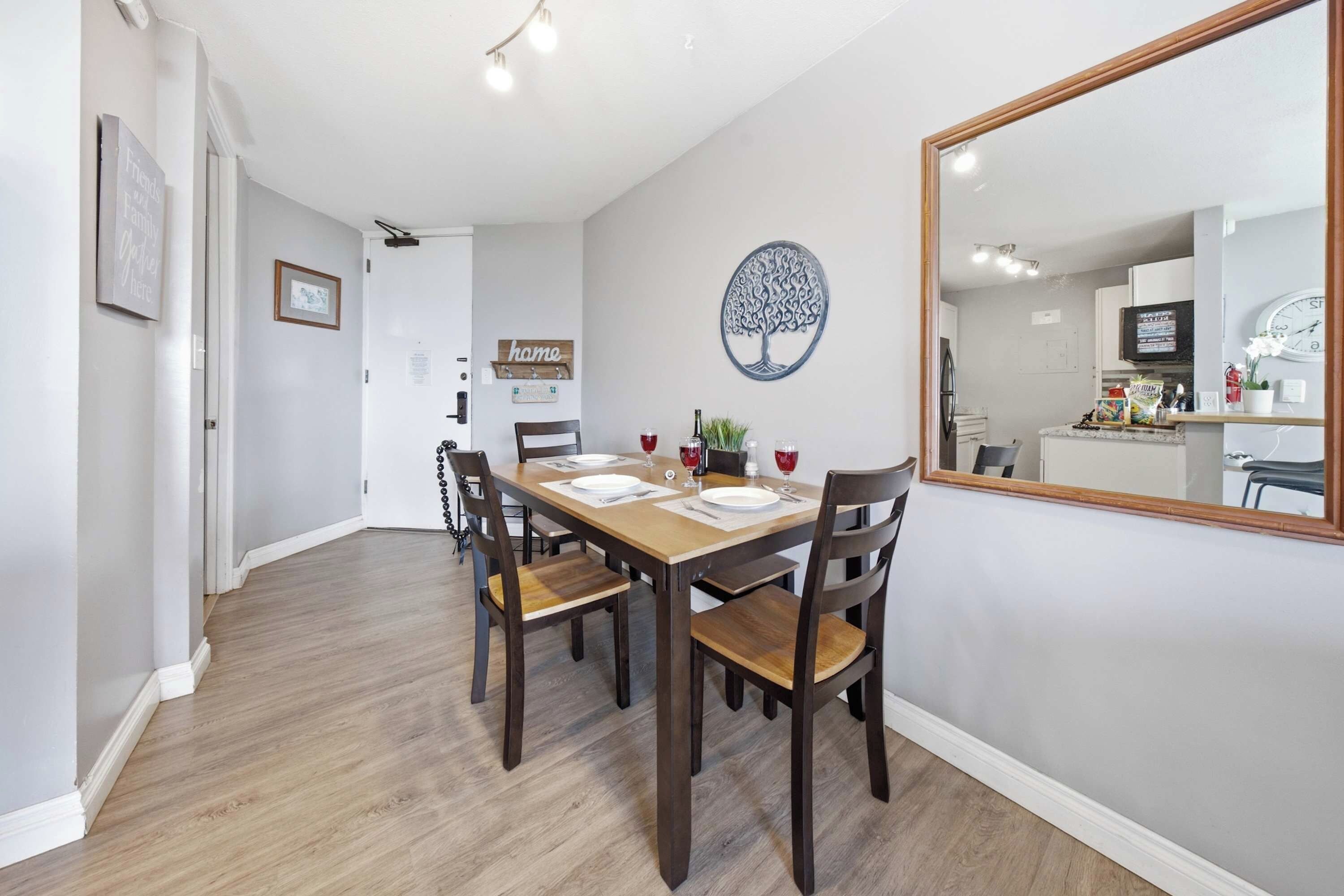 A dining area with a wooden table set for four, surrounded by chairs, a decorative tree wall art, and a large mirror reflecting the room.