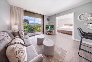 A modern hotel room with a balcony, a sofa set, a dining area, and a bedroom with a bed and a desk. - New Oceanfront Kihei Condo with Pool and Hot Tub (Kihei)