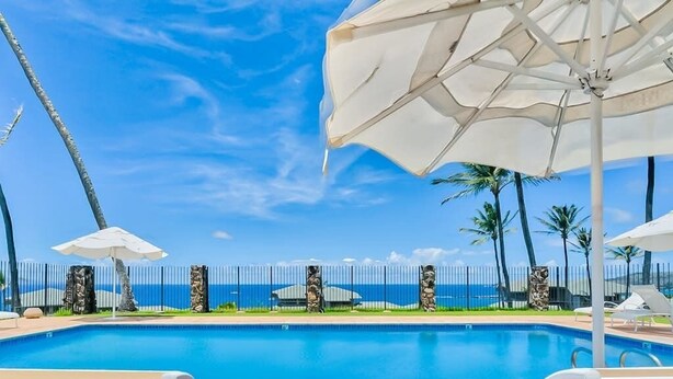 Ein Pool mit Liegestühlen und Sonnenschirmen, ein Zaun mit dekorativen Säulen sowie Blick aufs Meer.